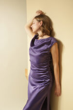 ZUMO FASHION satenska haljina rađena po meri sa blago prekrivenim ramenima, naborima koji oblikuju figuru i dubokim šlicem iznad kolena. Dostupna u četiri dužine, savršena je za svečane prilike i letnje proslave.